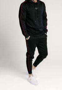 Felpa nera con strisce laterali rosse e logo, abbinata a pantaloni jogger neri con strisce simili. Scarpe da ginnastica bianche completano l'outfit.