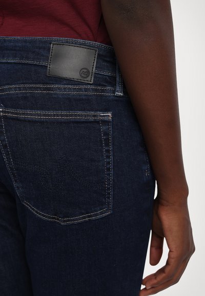 Dark blue denim jeans feature a leather patch on the waistband, contrast stitching, and two back pockets with subtle embroidery detail.