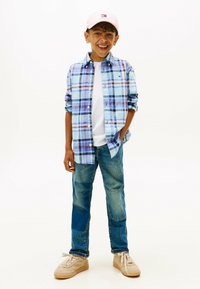 Blauw-wit geruite button-up shirt over een wit t-shirt, gecombineerd met blauwe jeans en beige sneakers. Roze pet met logo-details.