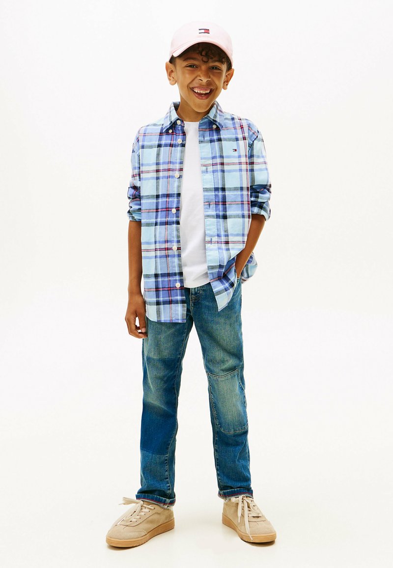 Blauw-wit geruite button-up shirt over een wit t-shirt, gecombineerd met blauwe jeans en beige sneakers. Roze pet met logo-details.