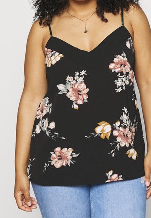 Femme portant un haut sans manches noir à fleurs roses et jaunes et un jean bleu, collier avec un petit pendentif rond visible.