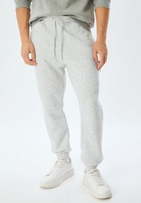 Grå bomulds sweatpants med elastisk talje og snor. Manchetter ved anklerne. Sammen med hvide sneakers. Glat, meleret tekstur.