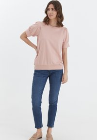 Helles pinkes kurzärmeliges Oberteil mit puffierten Ärmeln, Rundhalsausschnitt und geripptem Saum, kombiniert mit dunkelblauen, figurbetonten Jeans und beigen Slippern.