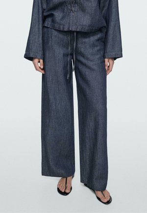 Personne portant un pantalon large en denim foncé et un haut à manches longues assorti avec des sandales noires à lanières sur un fond clair.