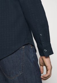 Námořnická modrá texturovaná košile s dlouhým rukávem a zapínacími manžetami, v kombinaci s tmavými džínami. Představuje jemný pruhovaný vzor a boční rozparek.