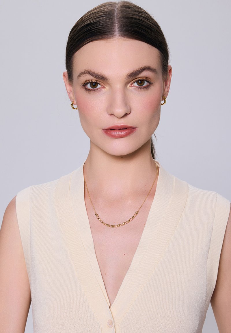 Bezramienna, jasnobeżowa bluzka o prążkowanej fakturze i głębokim dekolcie w serek, w połączeniu z delikatnym złotym naszyjnikiem oraz pasującymi kolczykami w kształcie kół.