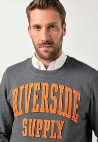 Uomo con capelli corti e marroni e barba, indossa una felpa grigia con la scritta arancione "RIVERSIDE SUPPLY" sopra una camicia bianca con colletto.