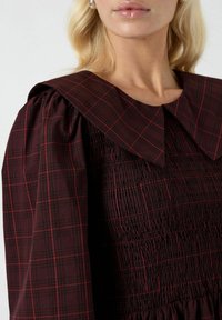 Abito a scacchi color bordeaux con colletto grande, caratterizzato da una texture plissettata e un design con arricciature. Maniche lunghe con dettagli arricciati.