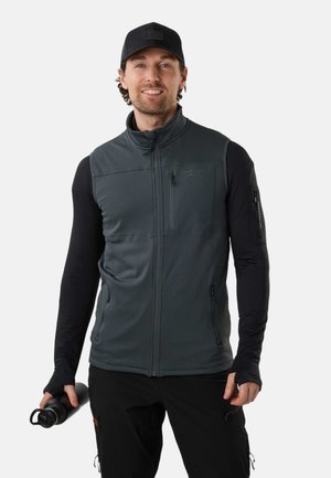 Mann trägt schwarze Kappe, dunkelgraue ärmellose Weste mit Reißverschluss über schwarzem Langarmshirt, hält schwarze Wasserflasche in der rechten Hand.