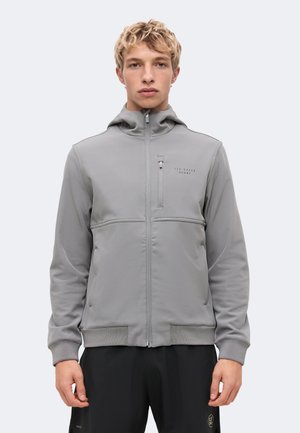 Un jeune homme pose avec une veste grise à capuche et à fermeture éclair, ornée d’un logo sur la poitrine, et un pantalon noir, sur un fond clair uni.