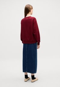 Felpa oversize bordeaux con polsini e orlo a costine, abbinata a una lunga gonna di denim blu e scarpe beige slip-on.