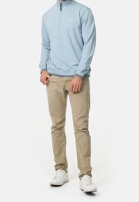 Portez un pull à fermeture éclair bleu clair avec des poignets et un ourlet côtelés, associé à un pantalon chino beige et des baskets blanches. Logo simple sur la poitrine.