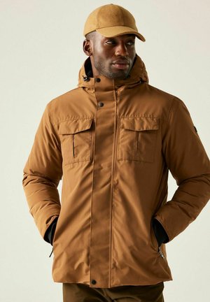 Mężczyzna ubrany w beżową kurtkę z kapturem, z zapiętymi kieszeniami na piersi oraz pasującą beżową czapką, z rękami w bocznych kieszeniach, patrzący na bok.