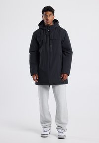 Black parka jacket with a front zip, hood, and chest pocket. Worn over grey sweatpants and white sneakers, featuring a relaxed fit and smooth texture.