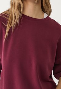 Bordeaux oversized sweatshirt met een gladde textuur, ronde halslijn en korte mouwen, gelaagd over een witte top, met minimale details.