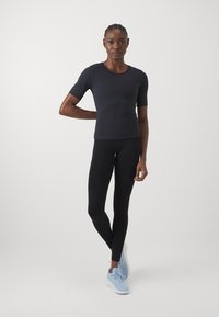 Sort kortærmet tætsiddende top parret med sorte leggings. Modellen bærer lyseblå træningssko. Enkelt design, glat tekstur.