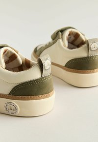 Børnesneakers i creme og olivengrøn, med en velcrorem, teksturerede detaljer og et løveansigt på hældappen.