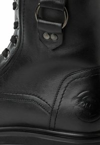 Primo piano di un boot in pelle nera che mostra la superficie texturizzata, occhielli in metallo con lacci neri, doppie borchie sulla cinghia e logo impresso vicino alla suola.