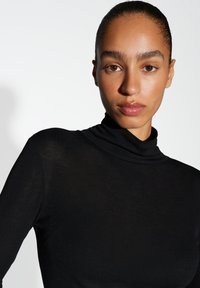 Femme aux cheveux tirés en arrière, portant un col roulé noir près du corps, regardant directement devant elle sur un fond clair. 