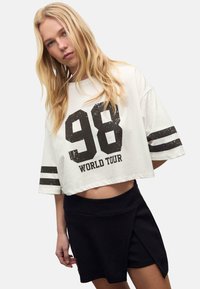 Witte cropped shirt met een versleten zwarte "98" en "WORLD TOUR" tekst, gecombineerd met een zwarte wikkelrok. Korte, losse mouwen.