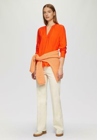 Blouse orange vif avec un col en V et des détails froncés, associée à un pantalon ample de couleur crème et un pull orange noué à la taille.