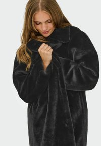 Donna con capelli lunghi che indossa e stringe un morbido cappotto in pelliccia sintetica nera con colletto alto, su uno sfondo chiaro e semplice.