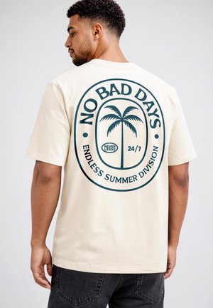 Mann, der von hinten zu sehen ist, trägt ein beigefarbenes T-Shirt mit großem Palmenmotiv und dem Text "No Bad Days, Endless Summer Division, 24/7."