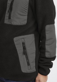 Schwarzer Fleece-Pullover mit grauen Nylonakzenten, ausgestattet mit mehreren Reißverschlusstaschen und texturiertem Stoff, hoher Kragen und lässige Passform.