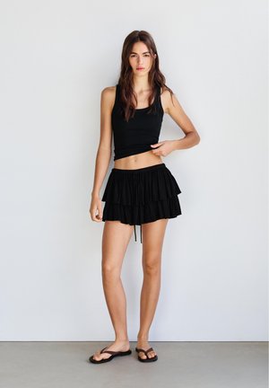 Młoda kobieta z długimi ciemnymi włosami, ubrana w czarny bezrękawnik typu crop top, czarną warstwową mini spódniczkę i czarne klapki, stoi przy białej ścianie.