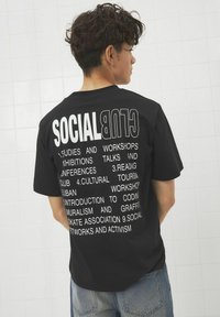 Mustra musta T-särk, mille seljal on suur valge tekst, mis loetleb "SOTSIAALKLUBI" ja töötoa teemad. Lühikesed varrukad ja vabalt langev lõige.