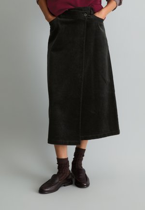 Persoon die een zwarte corduroy midi-rok met zakken, donkerbruine gerimpelde sokken en donkerbruine loafers draagt, staand tegen een effen lichte achtergrond.