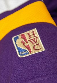 Lila Sporttrikotstoff mit gelben Akzenten, ausgestattet mit einem gestickten NBA-Logo-Patch, das den Text "HWC" und eine Spieler-Silhouette zeigt.