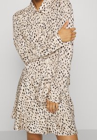 Béžové šaty košilového typu s černým tečkovaným vzorem, dlouhými rukávy, zapínáním na knoflíky a mírně rozšířeným lemem. Měkká, lehká látka.