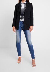 Svart skräddarsydd kavaj över en randig skjorta med knappar. Bärs med blå skinny jeans och vita klacksandaler. Slät textur och åtsittande design.