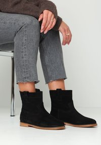 Black suede ankle boots with a flat tan sole, featuring a stitched design and pull tab, paired with cropped gray jeans.