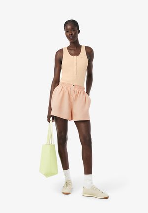 Femme portant un haut sans manches beige de style henley, un short rayé pêche, des chaussettes blanches, des baskets beiges, tenant un sac fourre-tout vert clair.