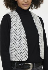 Strickweste mit Fischgrätenmuster in Weiß und Grau. Verfügt über eine weiche Textur und offenen Front, getragen über einem schwarzen Rollkragenpullover. Akzentuiert mit einem schwarzen Gürtel.
