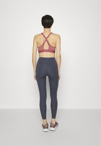 Reggiseno sportivo color marrone con spalline incrociate, design con schiena semi-aperta; abbinato a leggings alti in grigio scuro e scarpe da ginnastica viola.