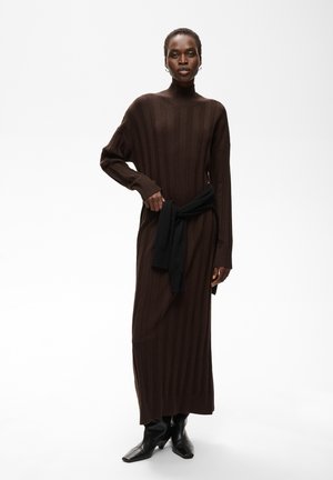 Femme portant une longue robe marron côtelée avec un pull noir noué autour de la taille et des bottes noires à talons sur un fond blanc.