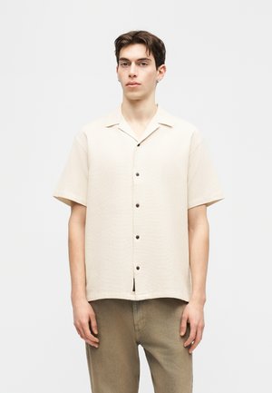 Young man wearing a short-sleeve beige button-up shirt and brown pants, standing against a plain light background.