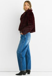 Burgundy faux fur jacket with a shawl collar, cropped design, and long sleeves, worn over blue straight-leg denim jeans and black heeled boots.