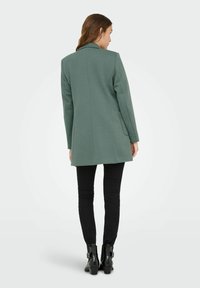 Femme portant un long blazer texturé vert, un pantalon slim noir et des bottines noires, se tenant de dos contre un fond uni.