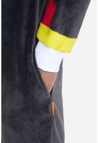 Tuta intera in fleece grigio carbone con tasca, caratterizzata da polsini a righe rosse, gialle e bianche. Include un'etichetta arancione vicino alla cucitura.