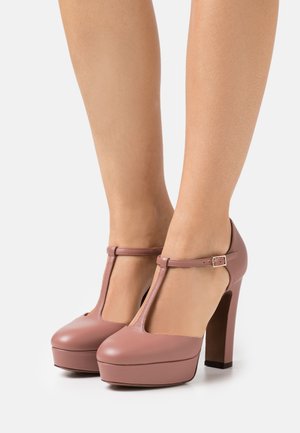 Vaaleanpunaiset korkokengät, joissa on T-muotoinen hihna, pyöreä kärki ja korotettu pohja. Nilkkahihnassa on solkilukko.