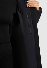 Persona che tiene aperto un cappotto nero mostrando la tasca interna con zip, indossando abbigliamento nero e su uno sfondo chiaro.