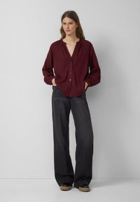 Burgundy button-up shirt with a loose fit, featuring a v-neck and gathered sleeves, paired with dark, wide-leg jeans and black platform shoes.