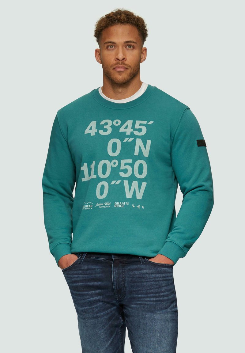 Sweatshirt de couleur sarcelle avec des coordonnées géographiques blanches imprimées sur le devant, col rond et manches longues. Porté avec un jean bleu foncé.