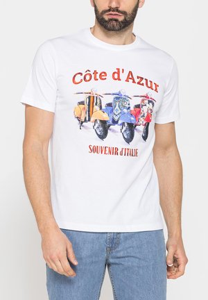 Uomo che indossa una t-shirt bianca con scooter colorati e testo "Côte d'Azur Souvenir d’Italie", abbinata a jeans azzurri chiari.