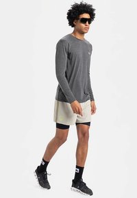 Dlhý sivý športový top s dlhým rukávom, béžové šortky s logom, čierne kompresné šortky pod nimi, čierne tenisky a slnečné okuliare.