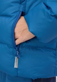 Blaues Puffer-Jackett mit einem Reißverschlussfach. Der Stoff ist glatt und hat einen subtilen Glanz. Die Hardware umfasst einen blauen Reißverschluss und ein Etikett mit "Mammut."